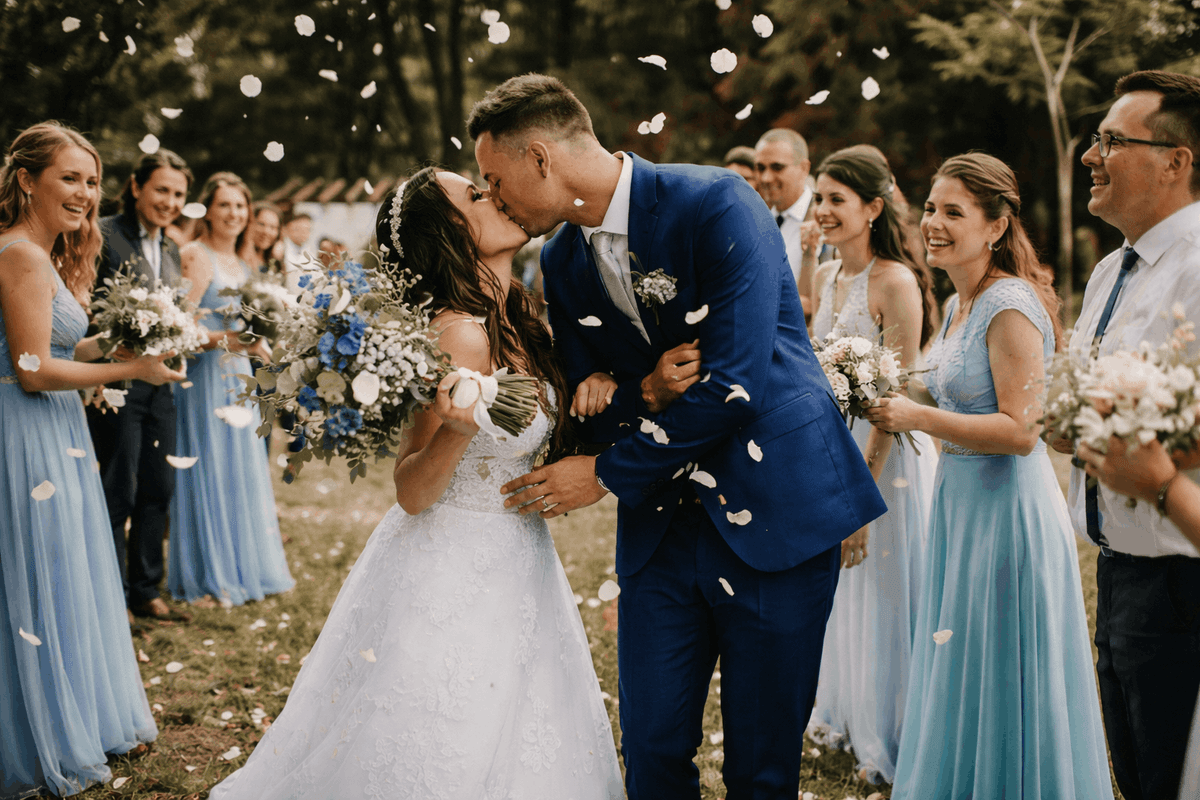 Brautpaar küsst sich nach der Trauung im Freien, während Gäste in hellblauen Kleidern Blumenblätter werfen.